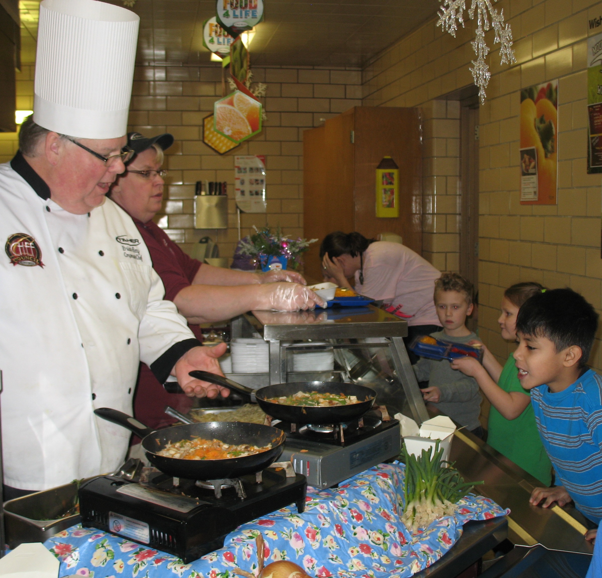 AS CHEF BRIAN prepares the chicken stir-fry meal on-site, kindergarten students file through the lunch line to pick up their lunch trays - including a very curious Tony Leyva. Behind him is Liliana Topete and Isaac Nowak. Serving up the meal to the right of Chef Brian is Carol Kipfer of Mountain Lake's Taher Food Service crew.